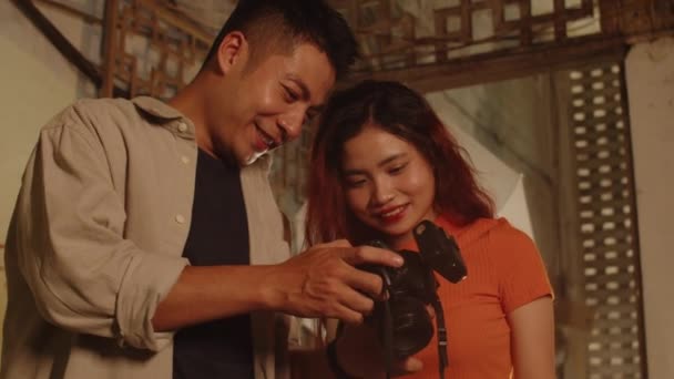 Bajo ángulo de joven pareja asiática comprobando fotos que hicieron en cámara dentro de edificio abandonado con paredes decoradas - Imágenes, Vídeo