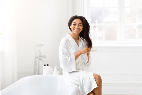 Tyyni afroamerikkalainen nainen kampaa hiuksensa tyytyväisellä hymyllä puhtaassa ja kirkkaassa kylpyhuoneessa - Valokuva, kuva