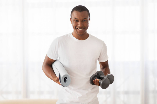 Mosolygós fekete férfi fehér pólóban, jóga szőnyeggel és súlyzókkal az edzéshez. - Fotó, kép