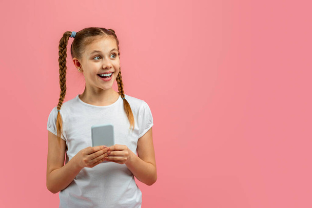 Excited young girl holding a smartphone looks away on a pink background showing curiosity and anticipation - Photo, Image
