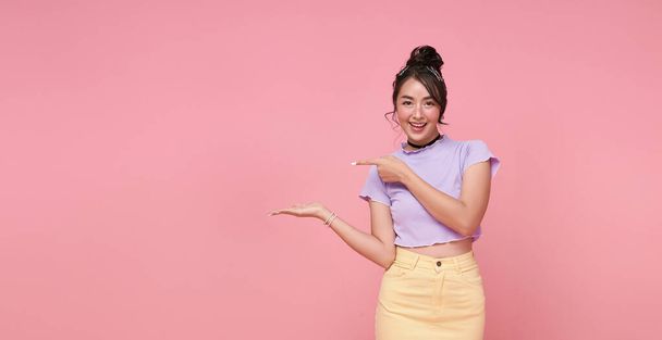 Hymyilevä nuori aasialainen teini tyttö etsii ja osoittaa sormella näyttää paikka mainonnan teksti Eristetty vaaleanpunainen tausta. - Valokuva, kuva