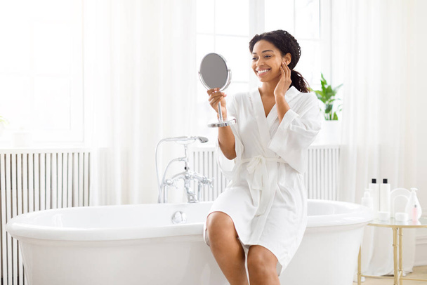 Cheerful African American lady in bathrobe applies skincare holding a mirror in a sunlit, modern bathroom - Foto, immagini
