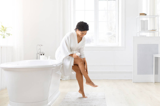 バスローブのアフリカ系アメリカ人女性は,セルフケアのルーチンを強調して,足の皮膚をチェックしていることが示されています - 写真・画像