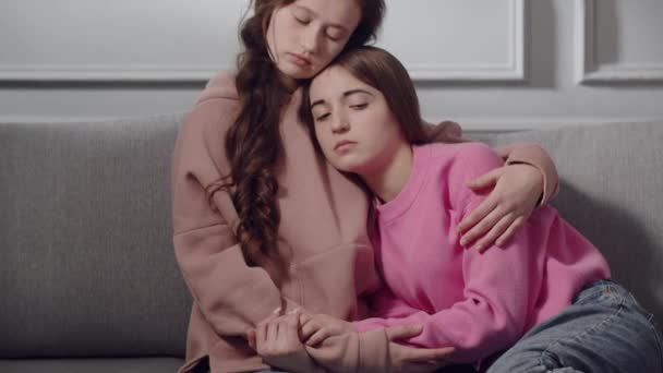 Zwei süße Teenager oder Studenten in traurigen Gefühlen sitzen auf dem Sofa, umarmen und trösten sich. Eine junge Frau blickt nach unten, stützt sich auf die Brust einer Freundin, die ihre Schulter streichelt, in einem schwierigen - Filmmaterial, Video