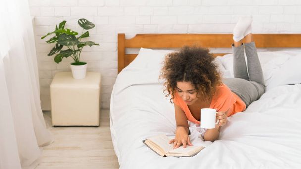 Афро-американская девушка читает книгу и пьет кофе в уютной спальне, пустое место
 - Фото, изображение
