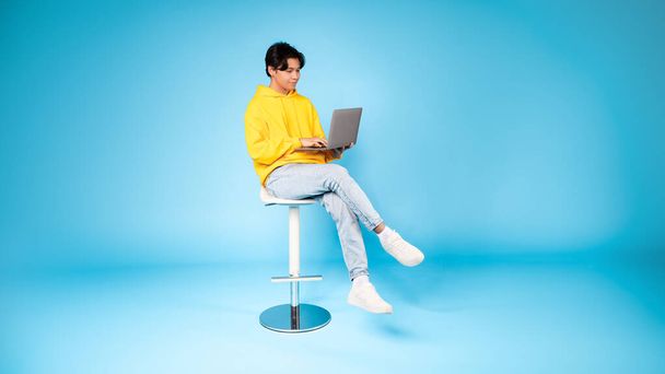 Tini ázsiai fiú ül egy magas széken egy laptop egy szemlélődő kifejezés ellen egységes kék háttér - Fotó, kép