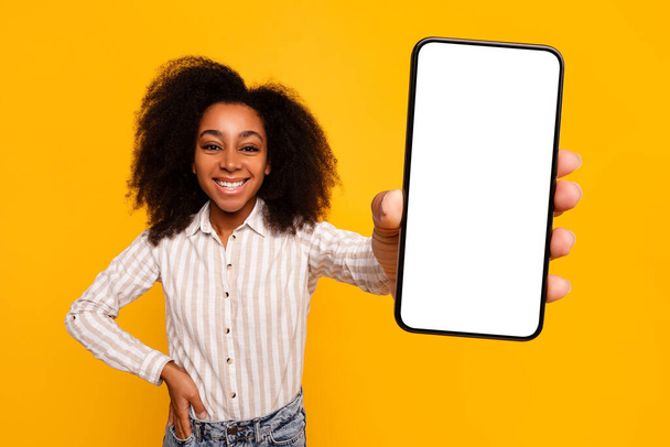 Joven mujer afroamericana alegre con el pelo rizado presenta una pantalla de teléfono inteligente en blanco, lo que sugiere la promoción de aplicaciones o sitios web - Foto, Imagen