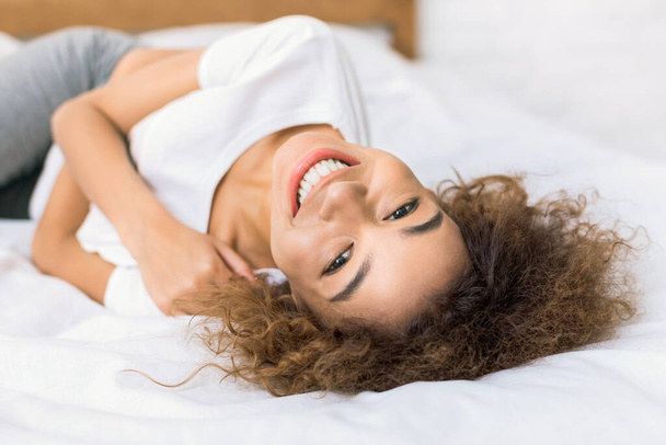 Mujer feliz acostada en la cama y mirando a la cámara, descansando en casa
 - Foto, Imagen