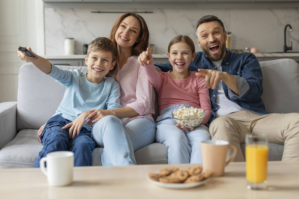 Egy család két gyerekkel izgatottan reagál valamire a TV-ben, miközben egy kanapén ülnek rágcsálnivalóval. - Fotó, kép