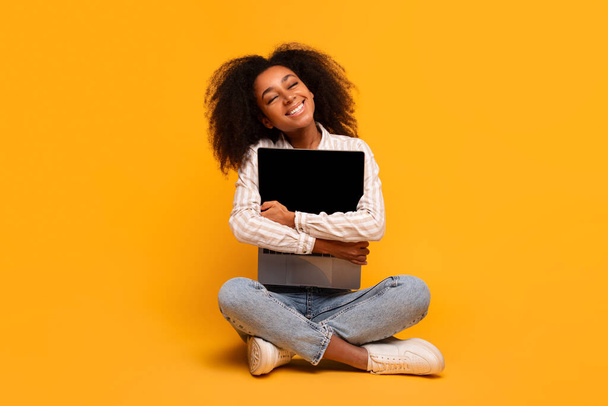 Młoda Afroamerykanka z kręconymi włosami siedząca na żółtym tle, przytulająca komputer z pustym ekranem - Zdjęcie, obraz