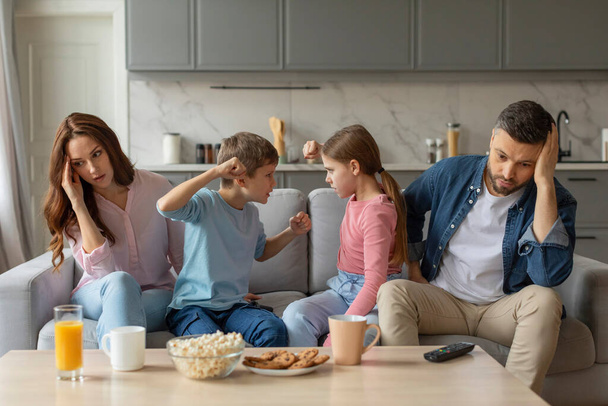 Fratelli ragazzo e ragazza cavillare su un gioco come i genitori si siedono indietro sensazione di stress e stanchezza in un soggiorno - Foto, immagini