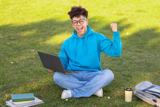 Radosny brazylijski student siedzący na trawie z laptopem, świętujący sukces, zeszyty i filiżankę kawy obok - Zdjęcie, obraz