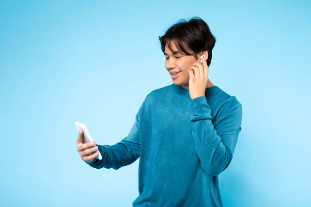Молодий азіатський хлопець насолоджується прослуховуванням музики або дзвінками за допомогою бездротових навушників і смартфона, синій фон - Фото, зображення