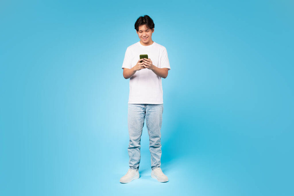 Junge Teenager asiatische Kerl tief in der Verwendung seines Smartphones vertieft, zeigt Technologie Engagement, blauer Hintergrund - Foto, Bild