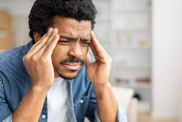 Zwarte gestresste man houdt zijn hoofd in pijn, toont ongemak van een ernstige hoofdpijn, thuis zitten en wrijven tempels, close-up met kopieerruimte - Foto, afbeelding