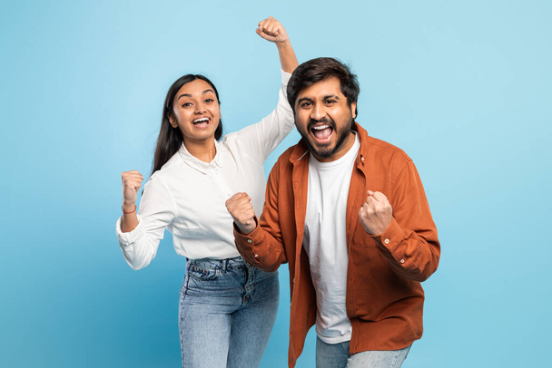 A joyful indian couple celebrates with fists raised in a victory or success gesture against a blue backdrop - Photo, Image