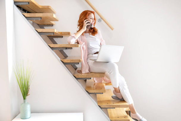 Feliz joven pelirroja trabajando en un portátil en su regazo, hablando por teléfono y usando camisa rosa, mientras está sentada en las escaleras en casa - Foto, imagen