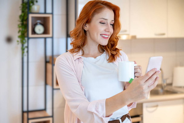 Portrait of attractive, young, ginger, red hair and smiling woman talking in a video call on her phone while drinking tea at home - Photo, Image