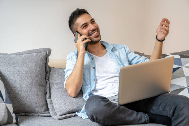 Portré fiatal, jóképű, mosolygós férfiról, aki telefonon beszél és használja a laptopját, miközben otthon ül a kanapén. - Fotó, kép