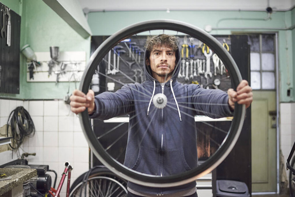 カメラを見ている若いヒスパニック系男性の肖像画は,自転車修理工場で自転車の車輪の縁を保持している. 選択的なフォーカスで構成される. 職場にいるリアルな人. - 写真・画像