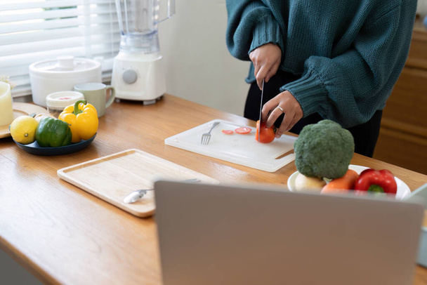 Eine Frau hackt pflanzliche Lebensmittel auf einer Arbeitsplatte aus Hartholz, während sie einem Rezept auf einem Laptop folgt. Die Geste des Obstschneidens zeigt ihre Kochkünste - Foto, Bild