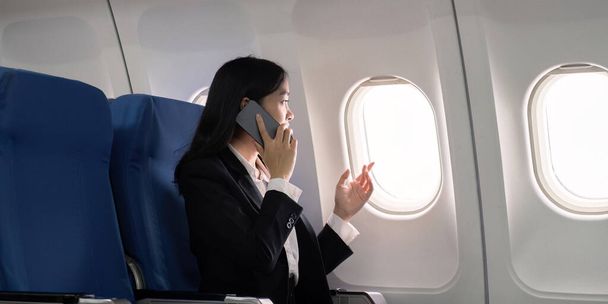 Azjatycka młoda bizneswoman sukcesu lub kobieta przedsiębiorca w formalnym garniturze w samolocie siedzieć w fotelu klasy biznes i używa smartfona podczas lotu. Koncepcja podróży i biznesu. - Zdjęcie, obraz