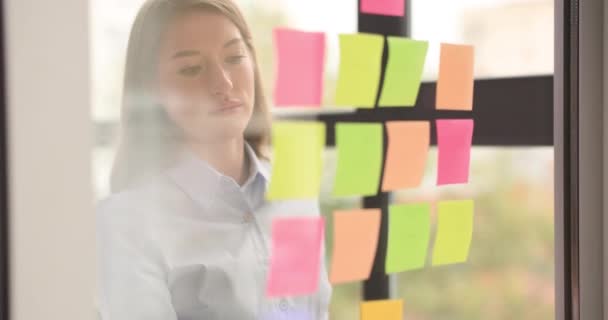 La empresaria reflexiva mira pegatinas de colores en una pared de vidrio y piensa en ideas de negocios. Planificación de la agenda y desarrollo de pequeñas empresas - Metraje, vídeo