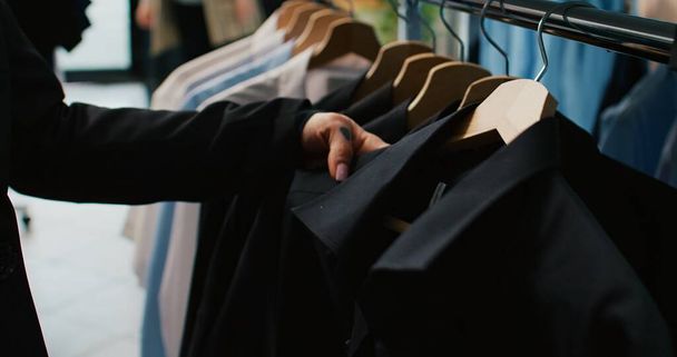 Eine elegante Frau begutachtet alle bunten Hemden, die im Bekleidungsgeschäft ausgestellt sind, und kauft nach trendigen Marken, um den Kleiderschrank zu erweitern. Der Kunde betrachtet Modeartikel, die auf Kleiderbügeln aufbewahrt werden. Aus nächster Nähe. Kamera B. - Foto, Bild