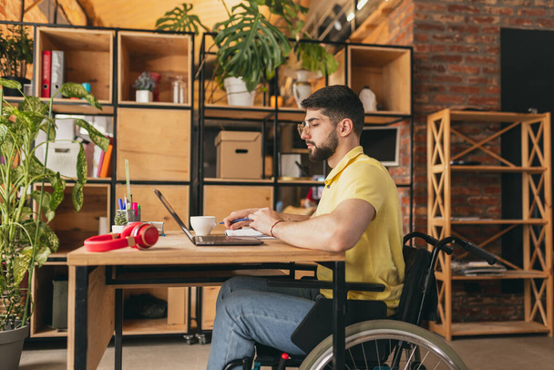 Young man, employee sitting on wheelchair and working remotely on laptop at home, Remote job, freelance. Concept of healthcare, lifestyle, wellness, comfort, empowerment - Foto, Imagen