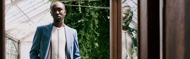 Un apuesto hombre afroamericano de sofisticado estilo elegante se encuentra frente a una ventana, enmarcada por un jardín verde vívido. - Foto, imagen