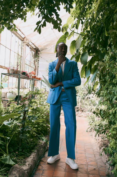 Hombre afroamericano guapo con un traje azul posando en un invernadero verde vivo. - Foto, imagen