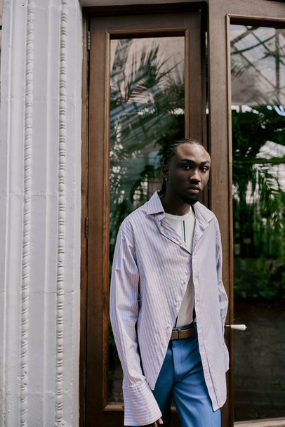 Όμορφος Αφροαμερικανός με κομψό στυλ στέκεται με αυτοπεποίθηση μπροστά από μια πόρτα σε ένα ζωντανό πράσινο κήπο. - Φωτογραφία, εικόνα