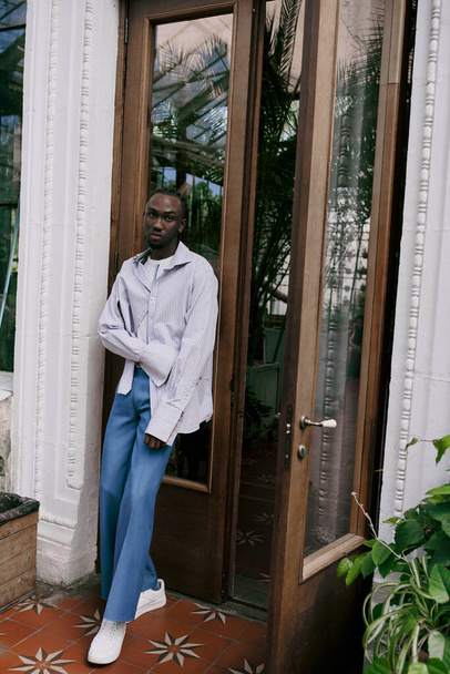Schöner afroamerikanischer Mann in elegantem Stil steht vor einer Glastür in einem lebhaften grünen Garten. - Foto, Bild