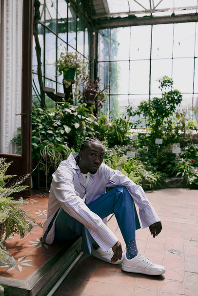 Handsome African American man in dapper style sitting on bench in vibrant greenhouse. - Photo, Image