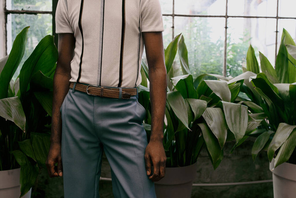 A stylish African American man poses elegantly among a variety of potted plants in a lush garden. - Photo, Image