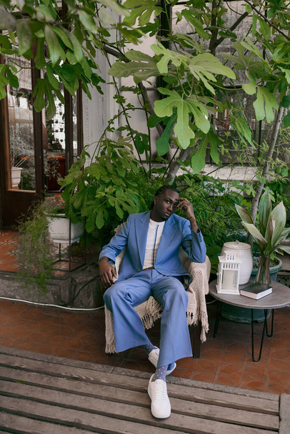 Ein gutaussehender afroamerikanischer Mann im blauen Anzug sitzt auf einer Bank in einem lebhaft grünen Garten. - Foto, Bild