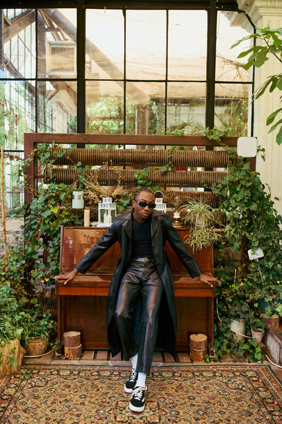 スタイリッシュなアフリカ系アメリカ人の男が活気に満ちた植物でいっぱいの部屋で優雅にピアノの上に座る. - 写真・画像
