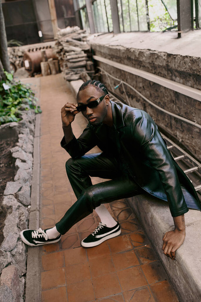 Handsome African American man in black leather jacket and sunglasses sitting on ledge. - Photo, Image
