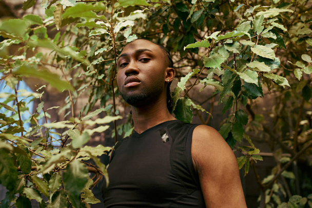 Ένας όμορφος Αφροαμερικάνος με κομψή ενδυμασία στέκεται περήφανα μπροστά από ένα καταπράσινο δέντρο σε έναν καταπράσινο κήπο. - Φωτογραφία, εικόνα