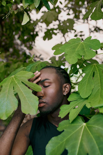 Un bel homme afro-américain les yeux fermés, caché derrière un arbre dans un jardin verdoyant. - Photo, image