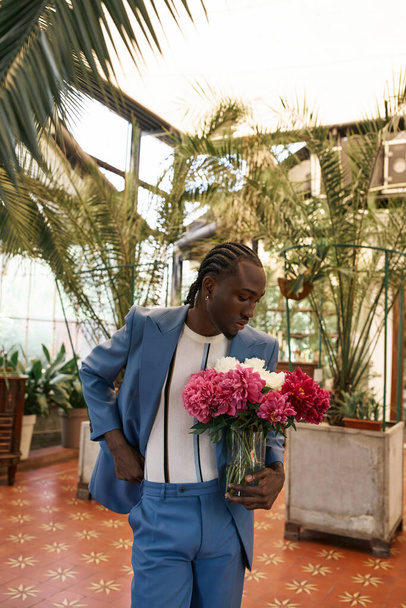 Anspruchsvoller afroamerikanischer Mann im blauen Anzug mit einem Strauß bunter Blumen in einem lebendigen grünen Garten. - Foto, Bild