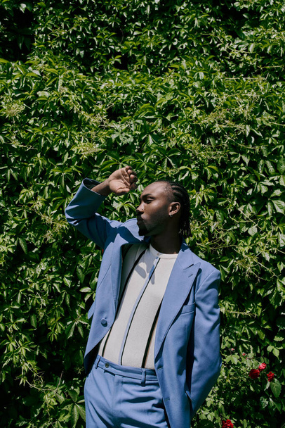 緑の茂みの前にエレガントに立っている青いスーツのスタイリッシュなアフリカ系アメリカ人男性. - 写真・画像