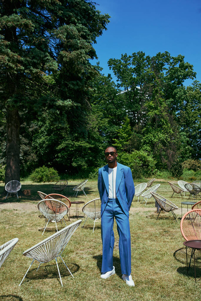 Schöner afroamerikanischer Mann im blauen Anzug inmitten von Stühlen im lebhaften grünen Garten. - Foto, Bild