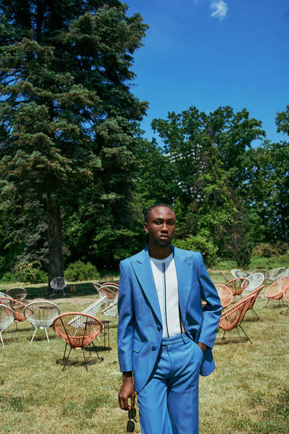 Un sofisticato uomo afroamericano in un elegante abito blu posa con grazia in un lussureggiante campo verde. - Foto, immagini
