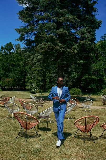 Przystojny mężczyzna w wyrafinowanym niebieskim garniturze stojący na polu krzeseł w żywym zielonym ogrodzie. - Zdjęcie, obraz