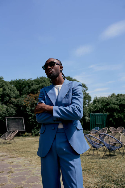 Sofisticato uomo afroamericano in abito blu e occhiali da sole che colpisce una posa in un vibrante giardino verde. - Foto, immagini