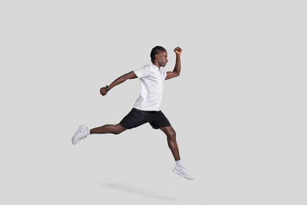 Imagen dinámica de un hombre afroamericano capturado en una pose de carrera en el aire sobre un fondo gris, que ilustra el movimiento y el atletismo - Foto, Imagen