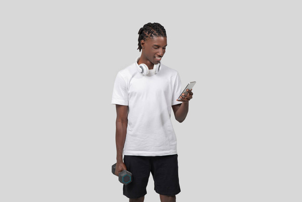 スポーツアフリカ系アメリカ人男性は,白い背景にダンベルを保持しながらスマートフォン画面を見ています - 写真・画像
