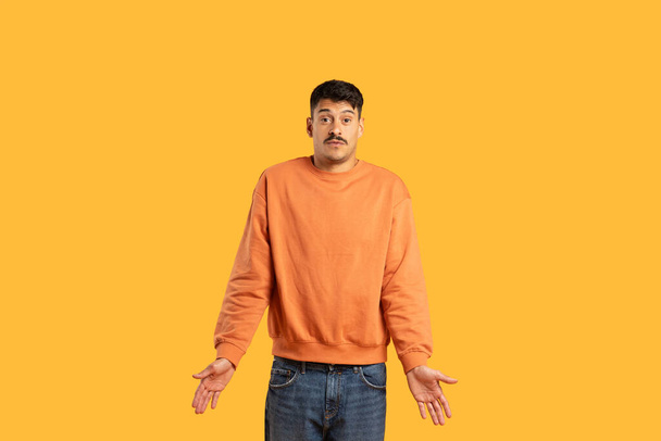 Zmatený muž s knírkem v oranžovém svetru pokrčí rameny s rukama otevřenýma na jasně žlutém pozadí - Fotografie, Obrázek