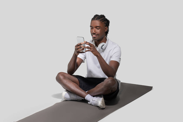 Un joven atlético afroamericano con un atuendo casual se sienta en una esterilla de yoga y envía mensajes en su teléfono inteligente en un entorno de estudio - Foto, Imagen